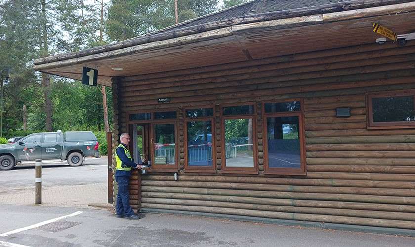 Security guard uses an intercom at a window labeled Security; building corner shows 1; surrounding context: log-cabin structure, parking lot, pickup truck, bollards, and trees.