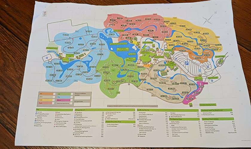 Folded theme-park map rests flat, displaying color-coded zones, rivers, paths, and icons with numerous tiny, unreadable labels; a legend and numbered lists span the bottom; set on a wooden table.