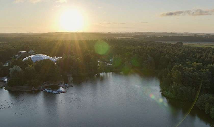 Sun sets, casting rays over a calm lake; surrounding pine forest hosts a marina with moored boats and a large domed building, seen from above in tranquil, rural landscape.