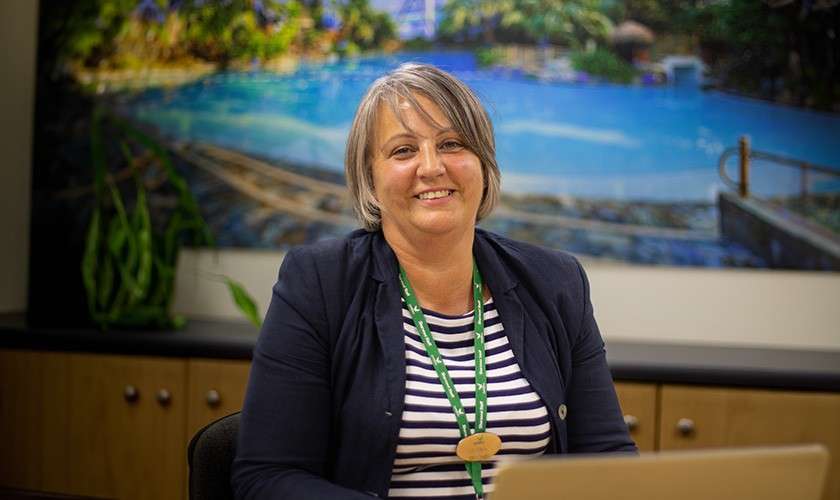 Person smiles while seated at a desk, working on a laptop; lanyard around neck. Behind, office cabinets and a large mural of a tropical pool scene.