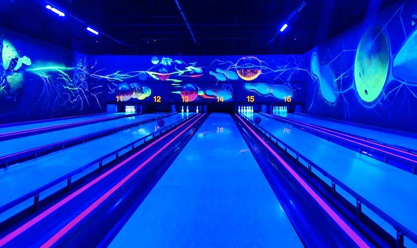 Bowling lanes glow, a ball rolls toward pins; surrounding walls show neon cosmic murals under blacklight. Lane markers read: 12 13 14 15 16.