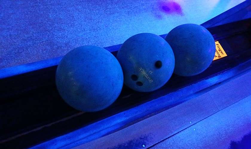 Three bowling balls rest in a return rack under blue neon lighting; the center ball shows finger holes. Visible text: “Brunswick 14” on the center ball and “Finger” on a yellow label.