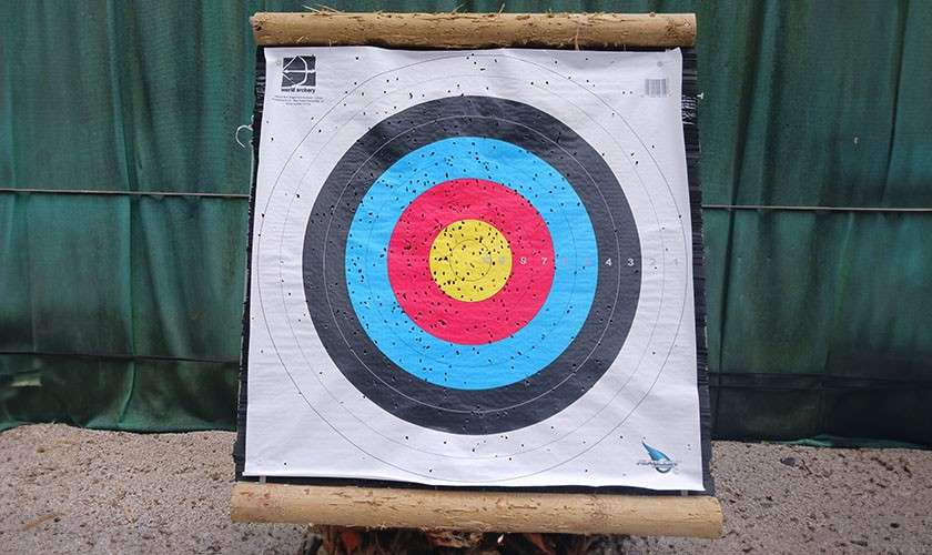 Archery target—riddled with arrow holes—stands on a wooden frame at an outdoor range with green curtain backdrop and gravel. Visible text: “world archery” and numbers “9 8 7 4 3.”
