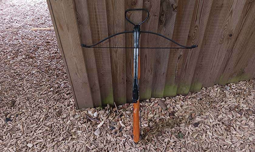Crossbow leans against a wooden fence, strung with slender metal limbs and a front foot stirrup, resting upright on woodchip-covered ground outdoors.