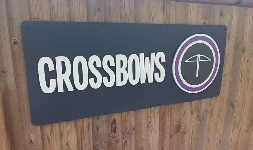 Sign displays "CROSSBOWS" and a crossbow icon, mounted on a wooden wall, indicating a crossbow activity or equipment area.
