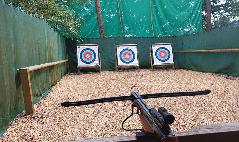 Crossbow aims forward toward three colorful bullseye targets at the far end of a woodchip-covered, netted outdoor range, with wooden rails and trees visible above the green screens.