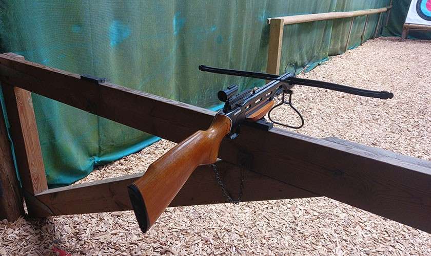 Crossbow rests on a wooden barrier, aimed downrange toward a target in the distance, within an indoor archery lane with green safety netting and a woodchip-covered floor.