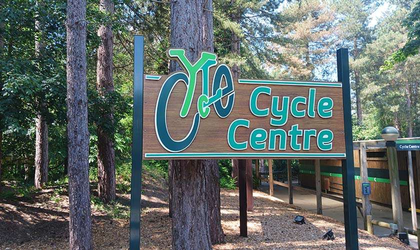 Wooden sign displays: “Cycle Centre.” In the background, a small building sign also reads: “Cycle Centre.” Surrounded by tall pine trees and a shaded path.