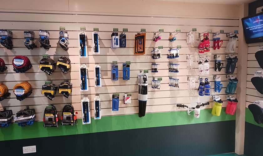 Cycling accessories hang neatly on a slatwall; coiled locks, pedals, pumps, bottle cages, and water bottles arranged in rows within a bike shop, green-trimmed walls and a small TV nearby.