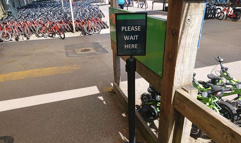 Sign reads "PLEASE WAIT HERE," mounted on a post by a wooden railing; situated in an outdoor bike rental area with clusters of parked bicycles on paved ground.