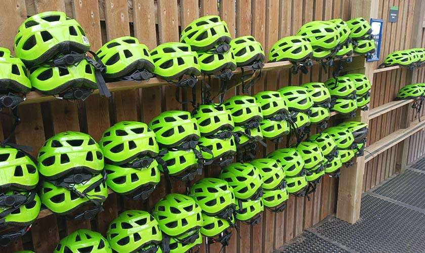 Neon-green bicycle helmets sit stacked in rows, hanging by straps, along wooden wall-mounted racks in a gear storage area with rubber floor mats.