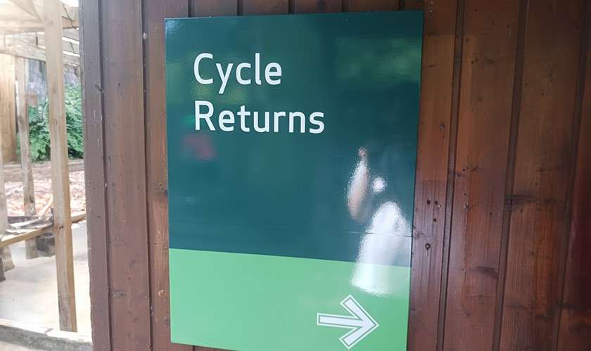 Sign directs cyclists right, mounted on a wooden wall at an outdoor facility. Text reads: “Cycle Returns” with a right-pointing arrow.