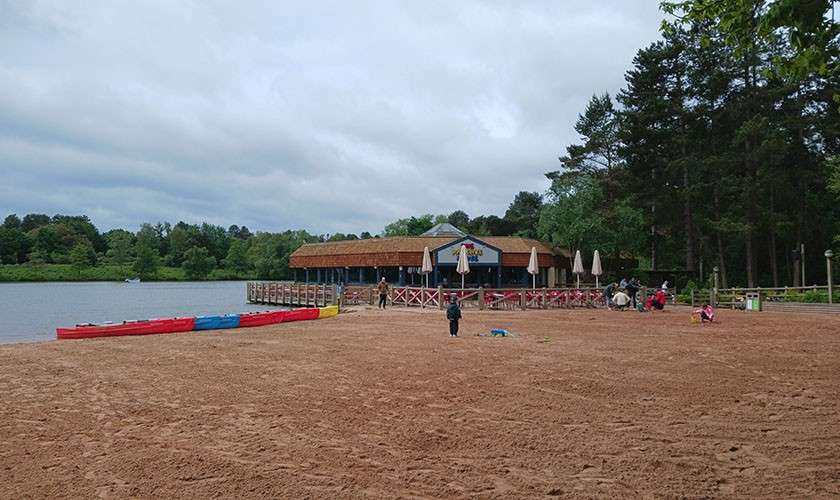 Beachgoers play on a sandy lakeshore, while kayaks lie lined up and others walk near a wooden pier and café with umbrellas; dense trees and cloudy sky frame the scene.
