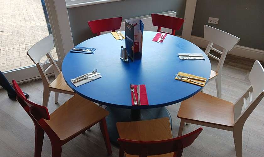 Round blue dining table—set with napkins and cutlery—stands indoors, six mismatched chairs around it; salt, pepper, and a menu holder centered, near a windowed door and wall outlet.