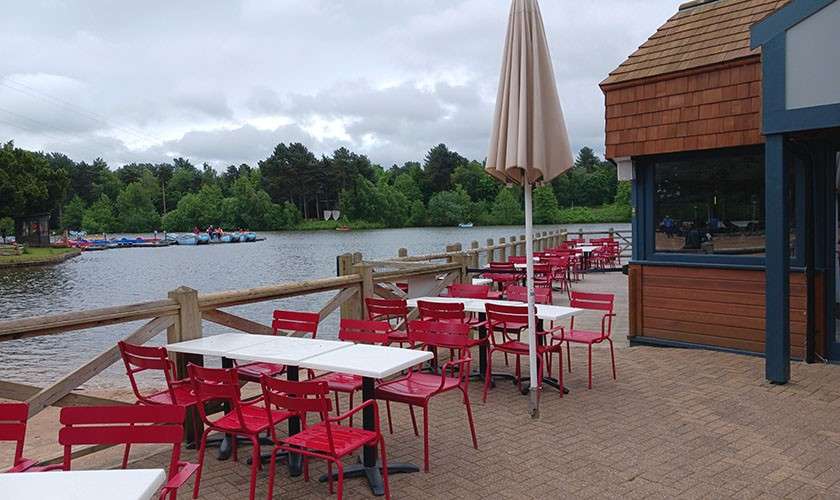 Café tables and red chairs sit empty under a closed umbrella, facing a lake. A wooden railing borders the terrace; paddle boats and trees line the opposite shore beneath clouds.
