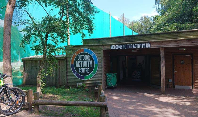 Wooden activity-center building displays signage, welcoming visitors; text reads: OUTDOOR ACTIVITY CENTRE and WELCOME TO THE ACTIVITY HQ. Surrounded by trees, fencing, and a bicycle near a low wooden barrier.