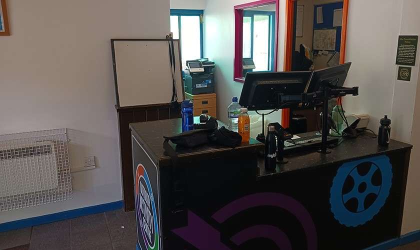 Reception counter holds dual monitors, keyboard, and bottles. Staff area sits behind with windowed booth, printer, and shelves. Nearby wall features blank whiteboard and caged heater in a compact office.