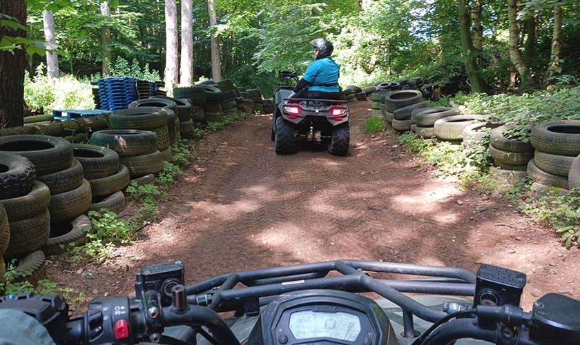 ATV riders navigate a dirt trail through a forest bordered by stacked tires; foreground shows handlebars, ahead a helmeted rider drives between trees under dappled sunlight.
