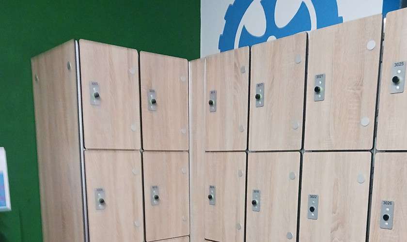 Lockers—closed—line a wall; each door has a knob and number plate. They stand against a green-and-white wall with a blue gear graphic. Visible numbers: 3025, 3026.