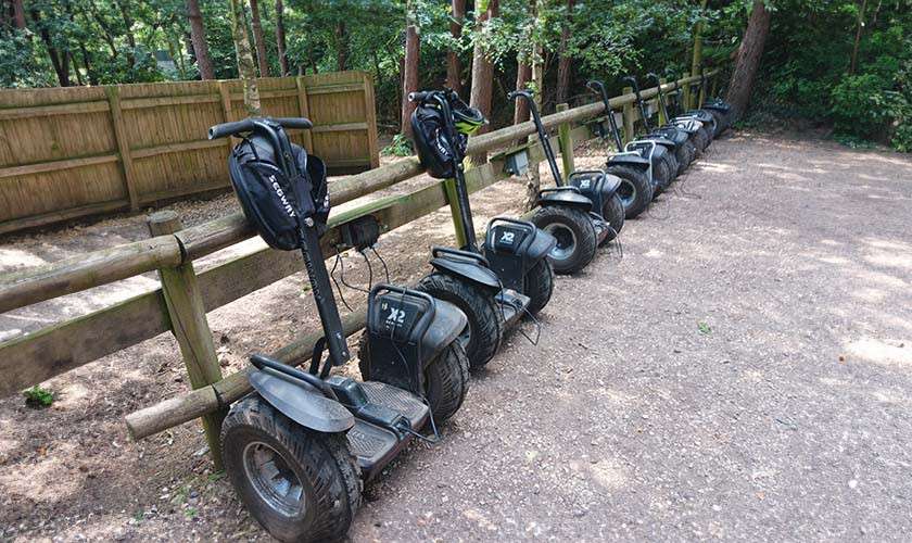 Row of electric scooters park leaning against a wooden fence on a gravel clearing in a forest; some have helmets on handlebars. Text: Segway, x2.
