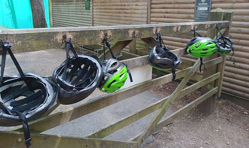 Five bicycle helmets hang by straps from hooks on a weathered wooden rack, outdoors beside a timber fence and gravel path, suggesting a bike rental or activity area.