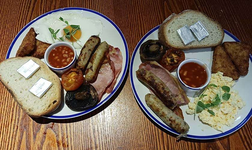 Two breakfast plates sit side by side, holding sausages, bacon, beans, eggs (fried and scrambled), toast with butter pats, grilled tomato and mushroom, and hash browns on a wooden table.
