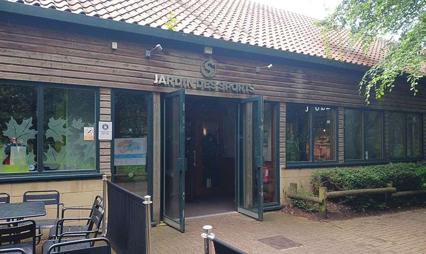 Double glass doors open, welcoming visitors to a wood-clad sports center; patio tables sit in front and trees surround the area. Text: JARDIN DES SPORTS.