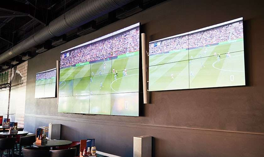 Wall-mounted video screens display a soccer match, showing a crowded stadium and pitch, while bar tables and menus sit below in a dim, industrial-style sports bar with exposed ducts.