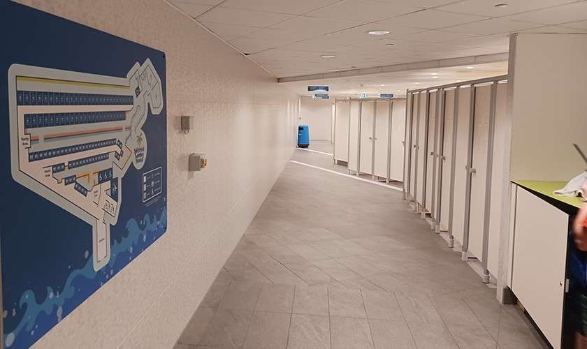 Row of bathroom stalls lines a long tiled hallway, leading toward a blue cleaning bin; a facility map hangs on the left wall under a low ceiling in a locker-room corridor.