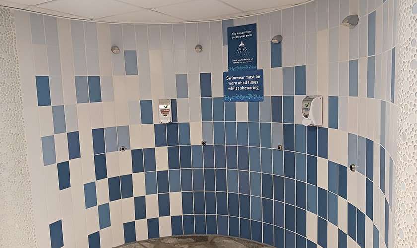 Curved tiled shower wall with multiple showerheads and soap dispensers in a pool changing area. Signs read: "You must shower before you swim" and "Swimwear must be worn at all times whilst showering."