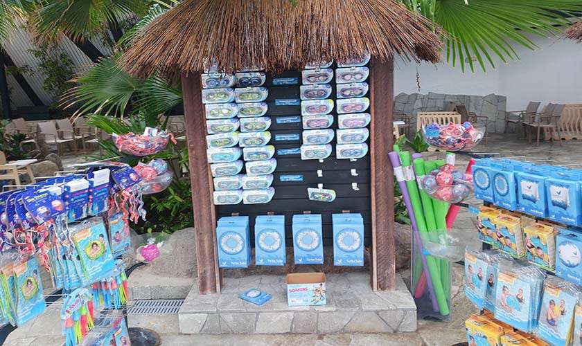 Pool-toy display showcases goggles, swim rings, and foam noodles, hanging on a thatched-roof kiosk, set within a tropical resort courtyard surrounded by palm plants, stone flooring, and nearby seating.