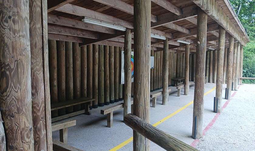 Wooden shelter provides seating, with log pillars supporting a roof and benches beneath. Fluorescent lights hang overhead; yellow and red boundary lines edge a gravel walkway amid surrounding greenery.