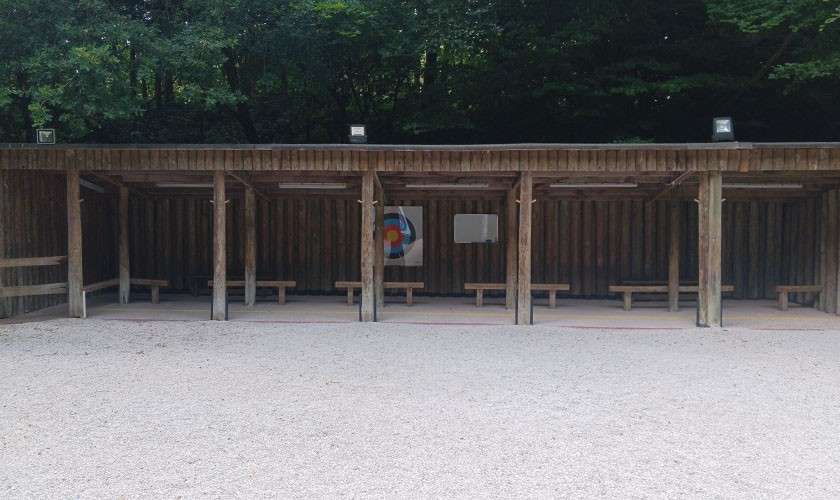 Archery range presents covered wooden shooting bays; a multicolored target hangs at center. Benches line each stall, while a gravel field stretches in front, bordered by dense trees above.