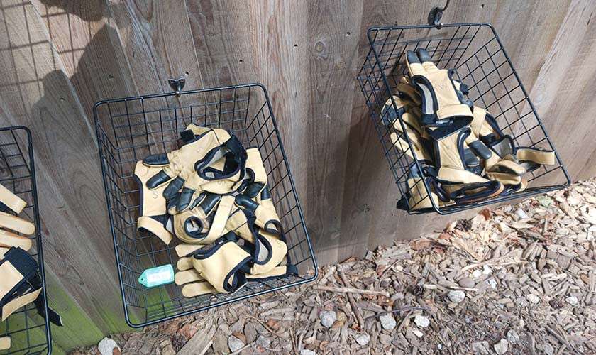 Wire baskets packed with tan-and-black work gloves hang from hooks, tilted outward, on a wooden fence beside a mulched area with small stones, suggesting organized outdoor storage.