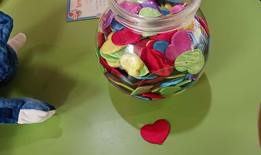 Glass jar holds many colorful satin heart-shaped confetti; one red heart rests on the green tabletop; a blue plush toy paw and a partially visible card are nearby.
