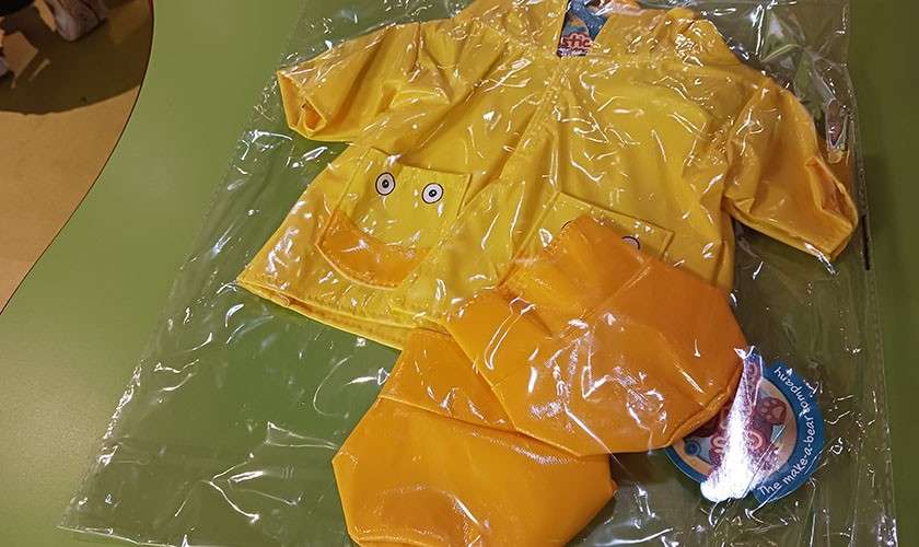 Child-sized yellow raincoat set rests sealed in clear plastic, featuring a duck-face pocket, with matching orange pieces, on a green table; attached tags present but text is unreadable.