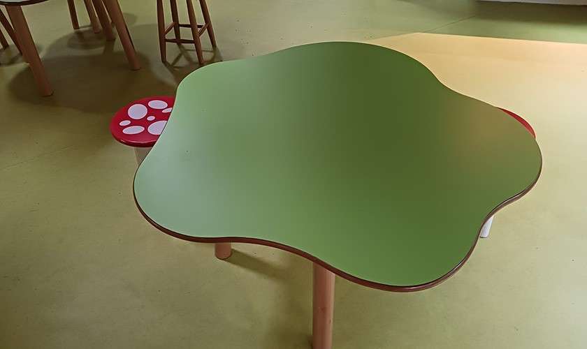 Green, wavy-edged children’s table sits on wooden legs, in a playroom/classroom, with small stools nearby, including a red seat dotted like a mushroom, on pale yellow flooring.