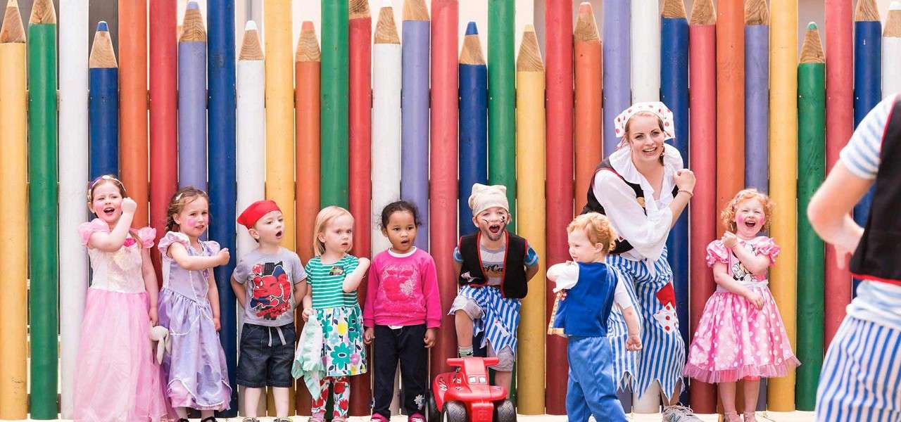 Children and a costumed adult laugh and pose, some wearing princess and pirate outfits; one rides a red toy car; backdrop features oversized vertical colored pencils forming a playful fence.
