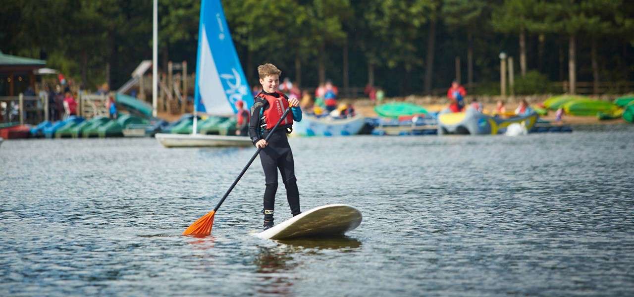 Child on stand-up paddleboard paddles forward with an orange oar; calm lake setting with sailboat, kayaks, and people near a wooded shore in the background. Visible text: Pico.