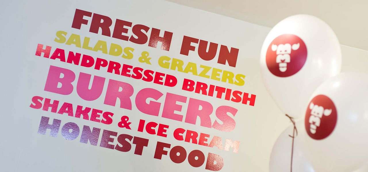 Colorful wall sign promotes menu while balloons float beside in a casual eatery. Text: FRESH FUN; SALADS & GRAZERS; HANDPRESSED BRITISH; BURGERS; SHAKES & ICE CREAM; HONEST FOOD.
