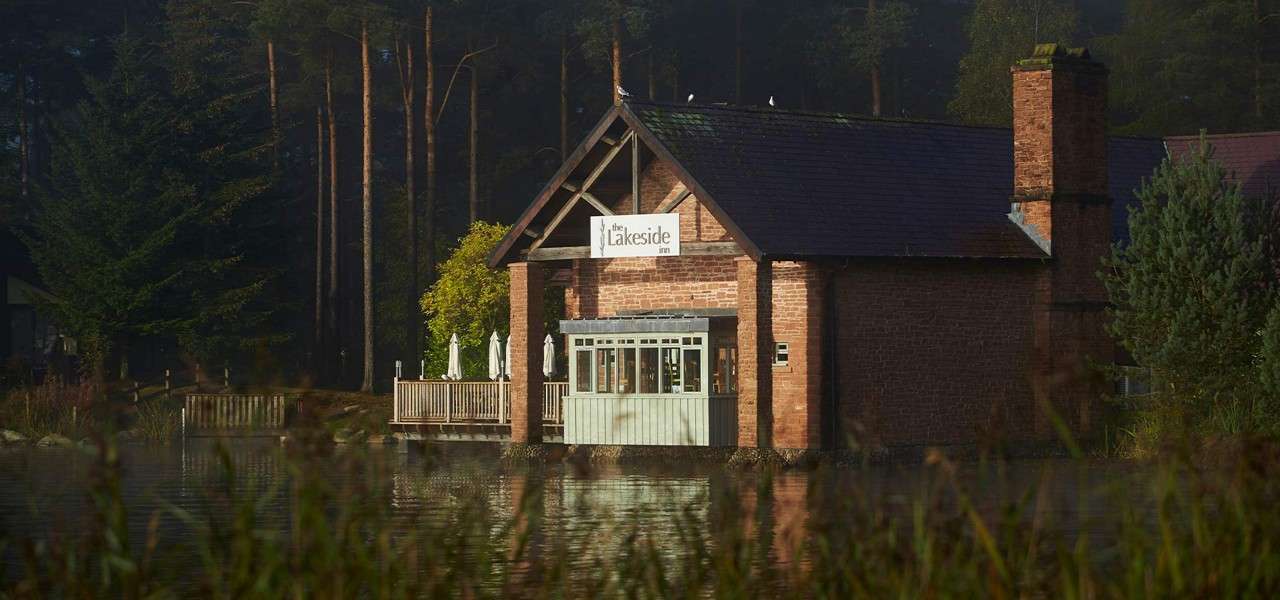 Brick lakeside restaurant sits beside calm water, displaying sign “the Lakeside”; umbrellas stand closed on a wooden deck, surrounded by tall pine trees and light morning mist.
