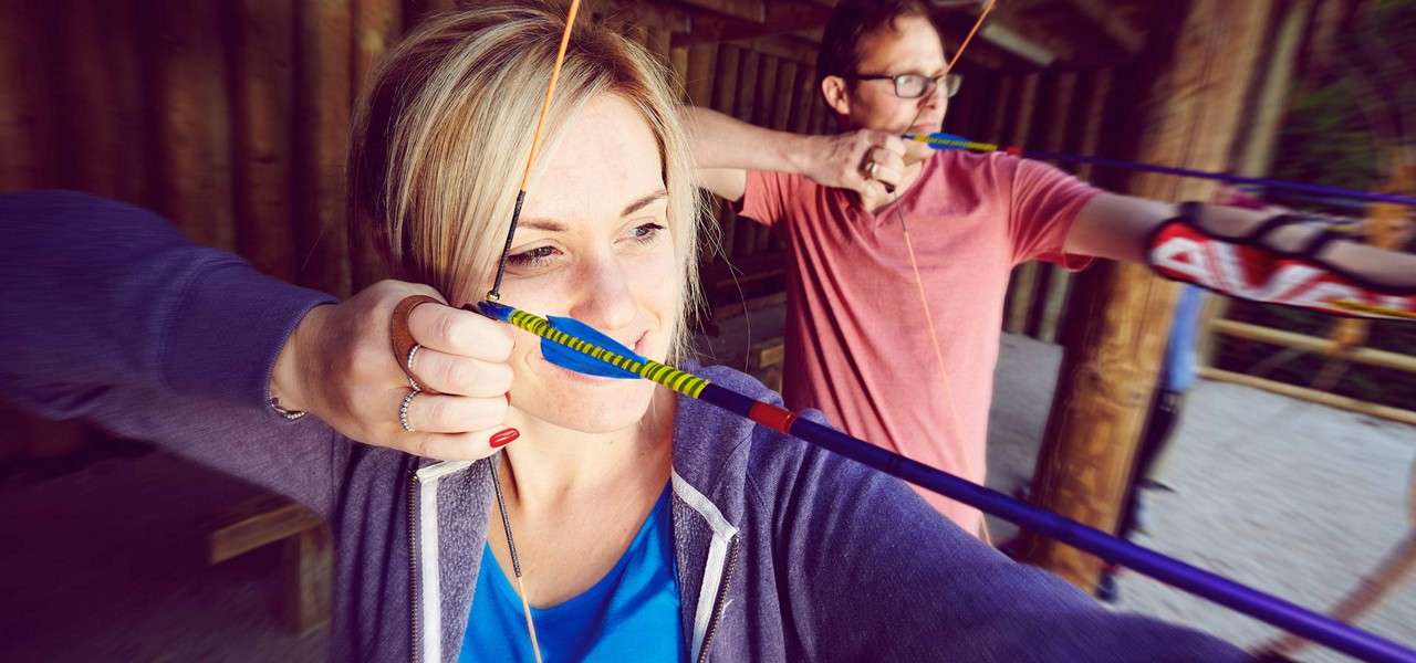 Two archers draw recurve bows, arrows nocked and strings pulled to cheek, focusing ahead. They stand side by side at a covered outdoor range with wooden posts and gravel.