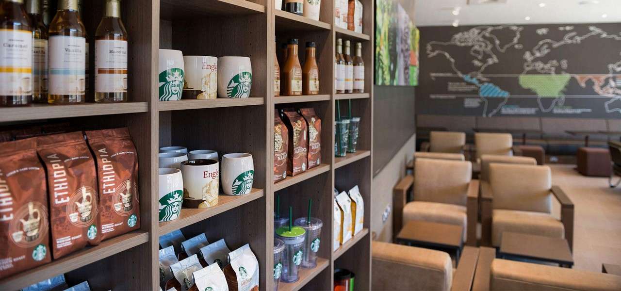 Merchandise shelves display coffee bags, mugs, syrups, and tumblers; a seating area lies beyond with beige armchairs and a wall map in a modern café. Text: ETHIOPIA, Caramel, Vanilla, Hazelnut.