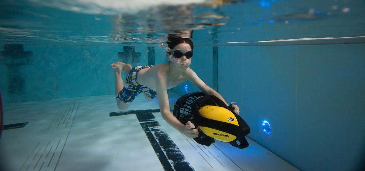 Child diver glides forward holding a yellow-and-black underwater scooter labeled "SEA-DOO," kicking in goggles; setting is a clear indoor swimming pool with lane markings and blue wall lights.