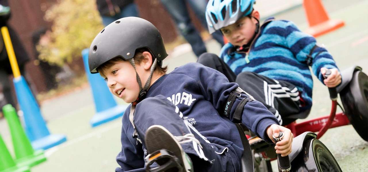 Two helmeted children steer low trikes, leaning and pedaling quickly, on an outdoor course lined with colorful cones. The foreground child smiles while gripping handles; the background child follows closely.