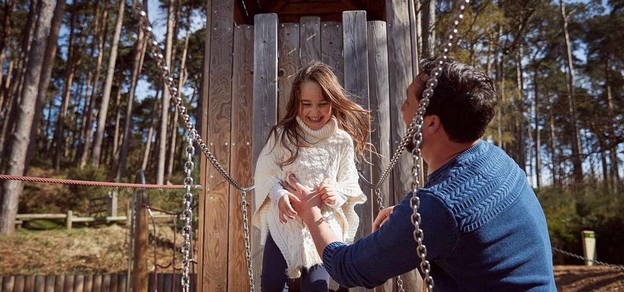 Girl steps down from a wooden play structure, smiling as an adult reaches to steady her; chains form handrails. In a forested playground, tall pines and fencing surround them.