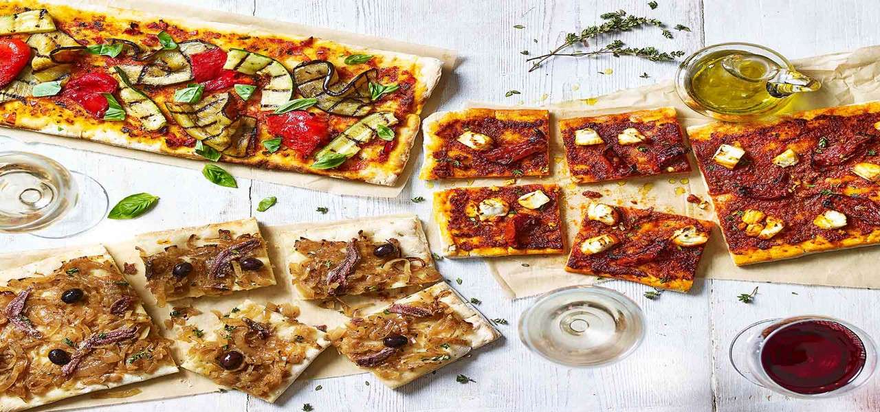 Rectangular pizzas rest sliced on parchment, topped with grilled vegetables, caramelized onions and anchovies, and sauced squares with cheese, beside olive oil, herbs, and glasses of wine on a table.