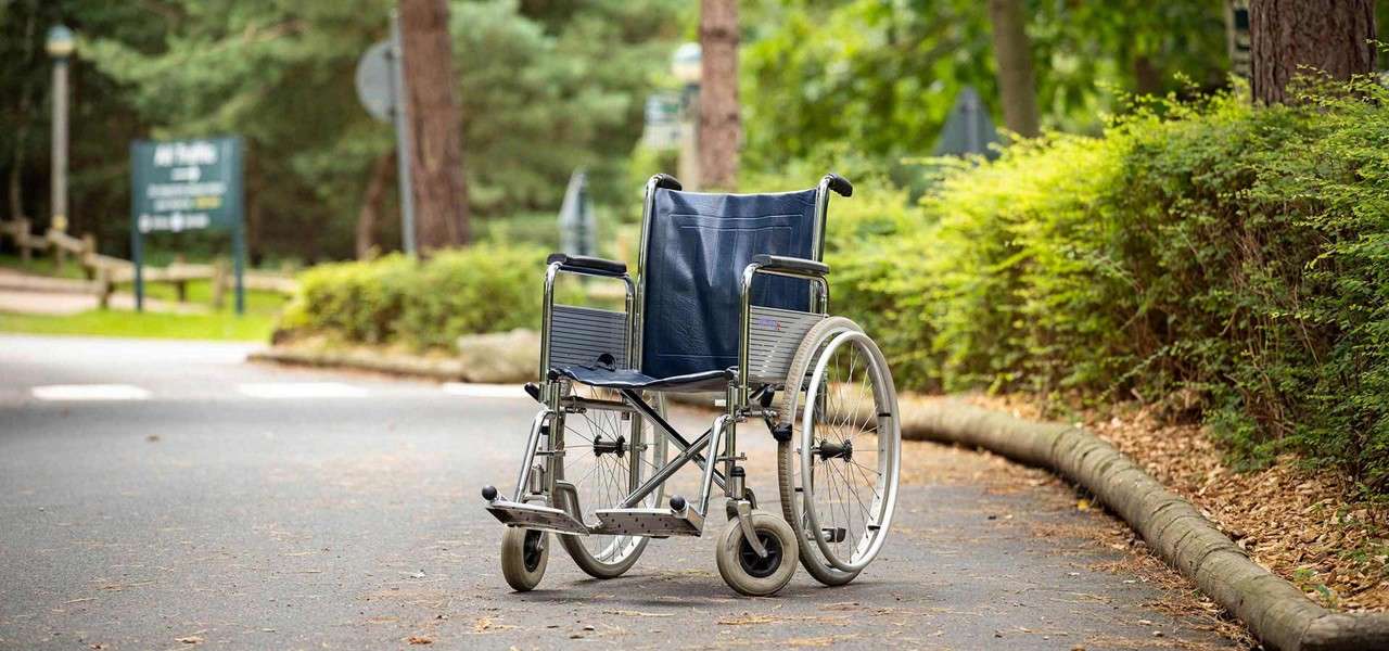 An empty manual wheelchair sits motionless on a paved path, its footrests down; surrounding context: leafy park with shrubs, trees, wooden edging, and blurred signposts in the background.