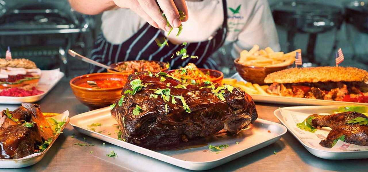 Roast beef is being sprinkled with chopped herbs, while a cook in an apron prepares dishes; surrounded by sides—fries, rice, sauces, sandwich, and grilled chicken—on a busy kitchen counter.