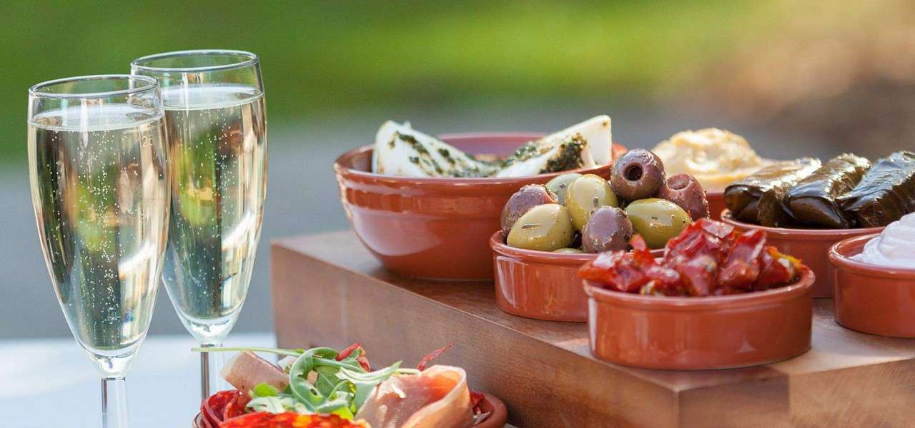 Tapas bowls present assorted olives, cheese, stuffed grape leaves, sun‑dried tomatoes, and dips, arranged on a wooden board; two flutes of sparkling wine stand beside them in a garden setting.